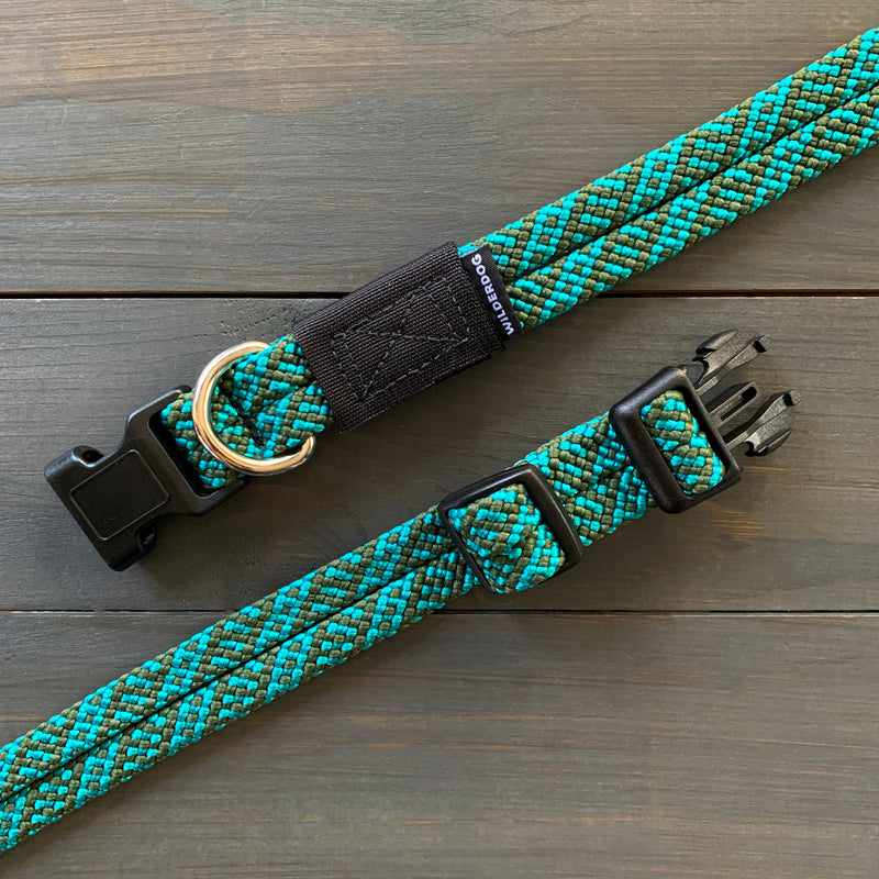 The Cascade Collar, a blue and green braided dog collar made from rugged rock climbing rope, includes a black plastic buckle and adjustment slider. Accompanied by a matching leash with a metal D-ring, it is showcased on a wooden surface.